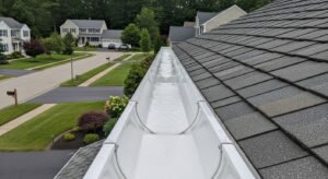 Gutter Services MA & RI: A close-up view of a clean white gutter system along a residential roof with gray shingles and a suburban neighborhood in the background.